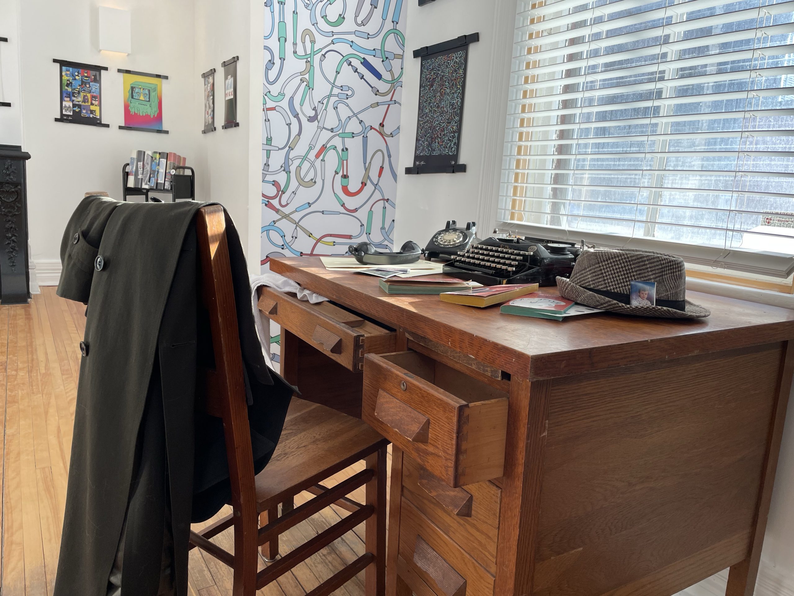 A type writer sits on a desk with a hat and a pen. An overcoat is thrown over a chair.