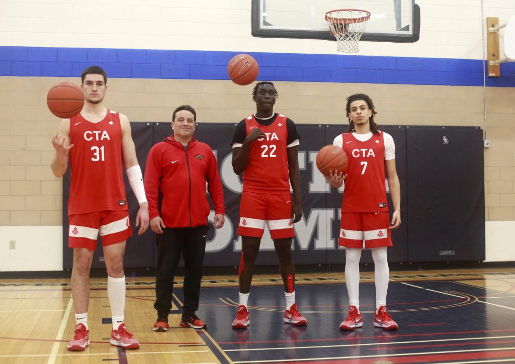 Four men wearing red pose for a photo. Three of them are spinning basketballs. They are standing in a gym. 