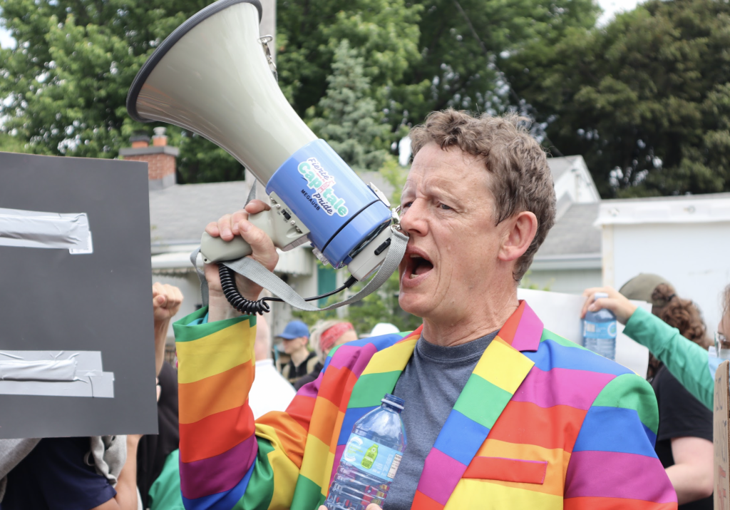 Ottawa Centre MPP Joel Harden chants into a megaphone. He’s wearing a rainbow suit. 