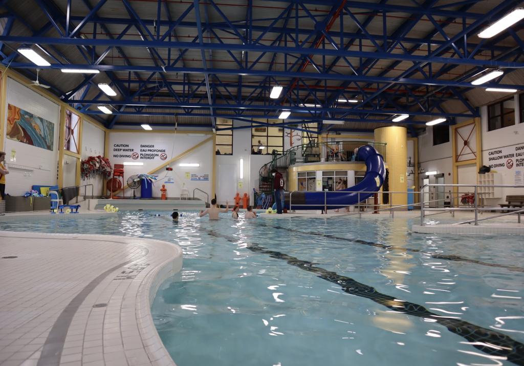 The pool in Dovercourt with a few people swimming in the background. A dark blue water slide is also in the distance.