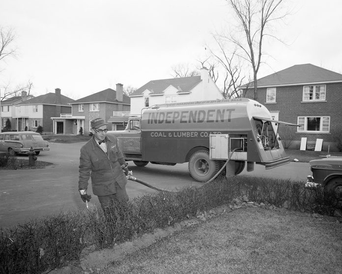 A black and white photo of a man taking a hose out of a coal and lumber truck. It’s taken in a residential neighbourhood