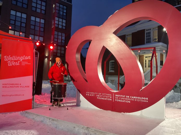 A woman plays a drum next to a very large red heart on display outside.