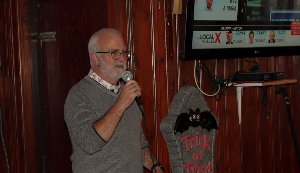 A man wearing gray sweater with a gray bears gives a speech