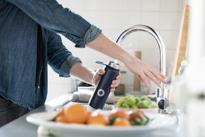 A person wearing a denim shirt holds a black water bottle under a silver tap. There are bowls of fruit next to the sink.