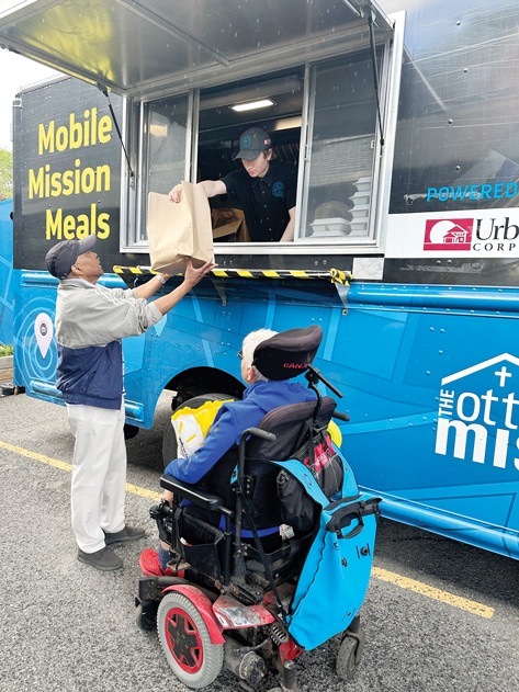 A man gets food from a food truck. A person in a wheelchair is waiting in line.