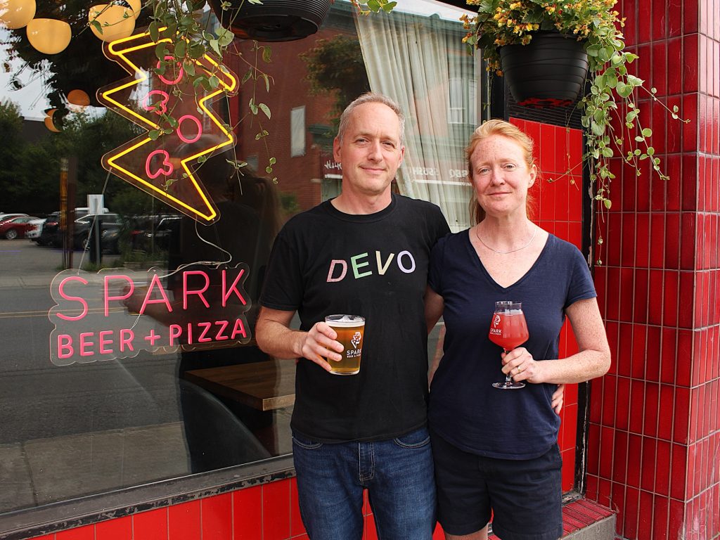 Two people stand outside if their storefront and hold glasses of beer.