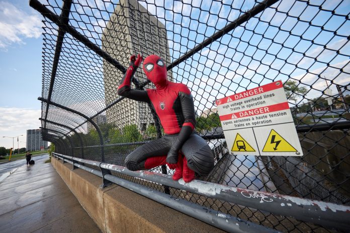 A man dressed up as Spider Man holds onto a railing near tunneys pasture. The LRT is seen in the distance.