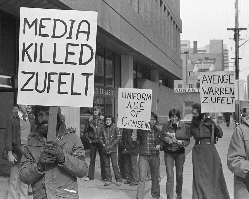 A black and white photo of demonstrators holding signs which read “Media called Zufelt”