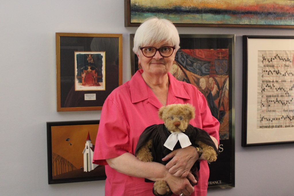 Tamra is wearing a pink shirt and poses for a photo holding a stuffed bear that is wearing a judges uniform.