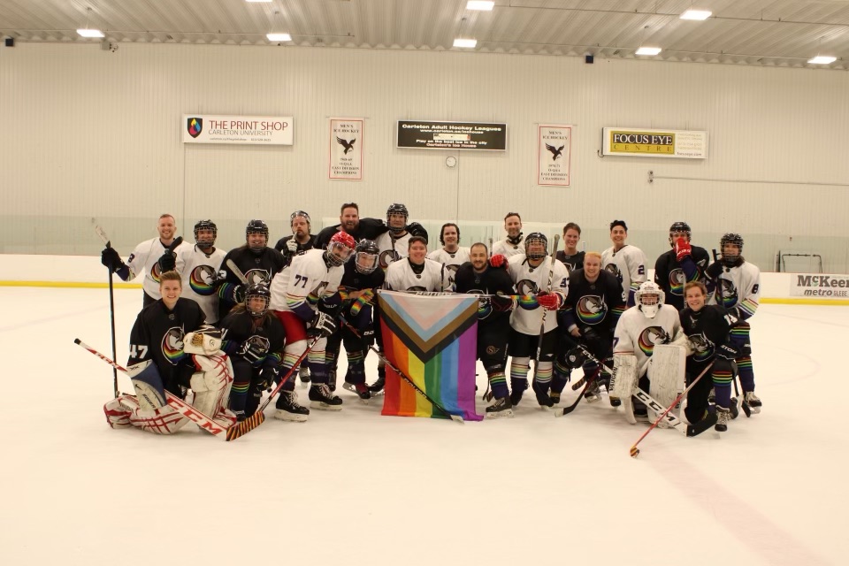 A group of hockey players pose for a photo on the ice while holding a pride flag. 