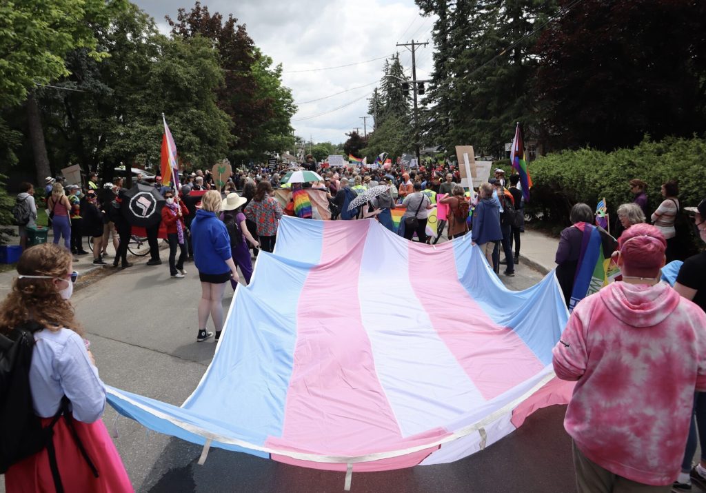 A large trans flag is carried down the street by multiple volunteers.