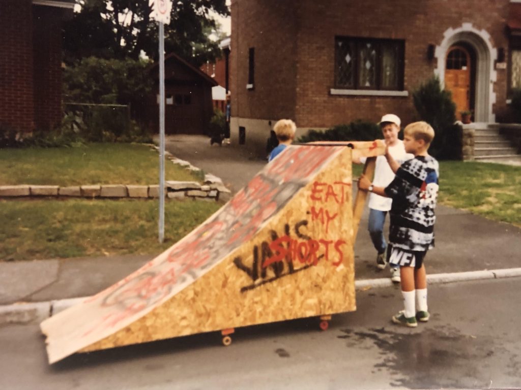 An old photo of theee young boys pushing a skateboard down a homemade wooden ramp.