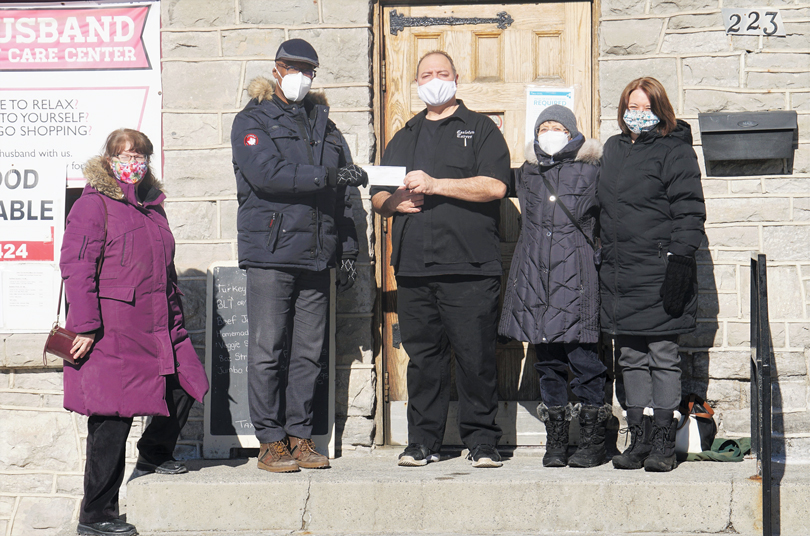 A group of five people stand on the steps of Carlton Tavern on a sunny day in winter. Two of them are holding a white cheque.