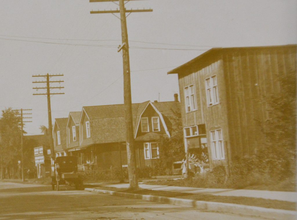 An old black and white photo of a residential street. A car is either driving through or parked.