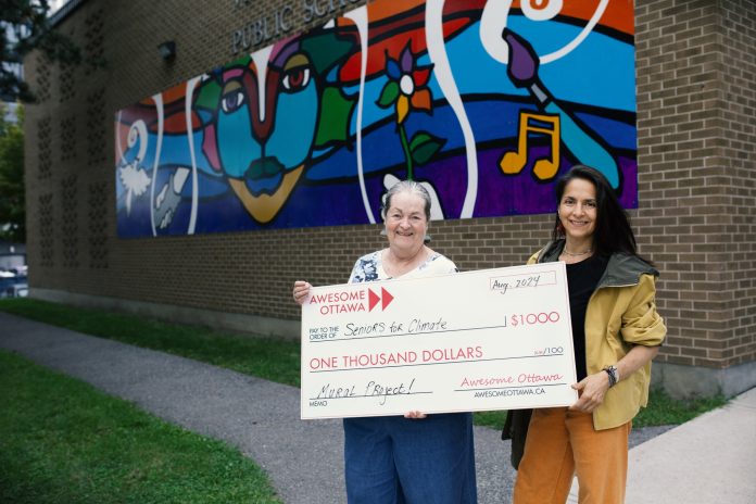 Two women stand outside a school that has a mural and holds a big cheque for $1000.